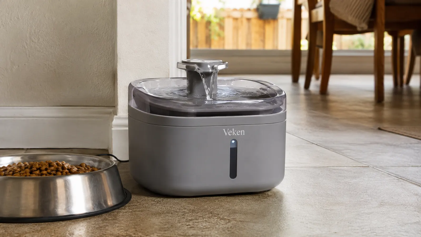 Grey plastic pet water fountain filled and running on a tile floor next to a food bowl